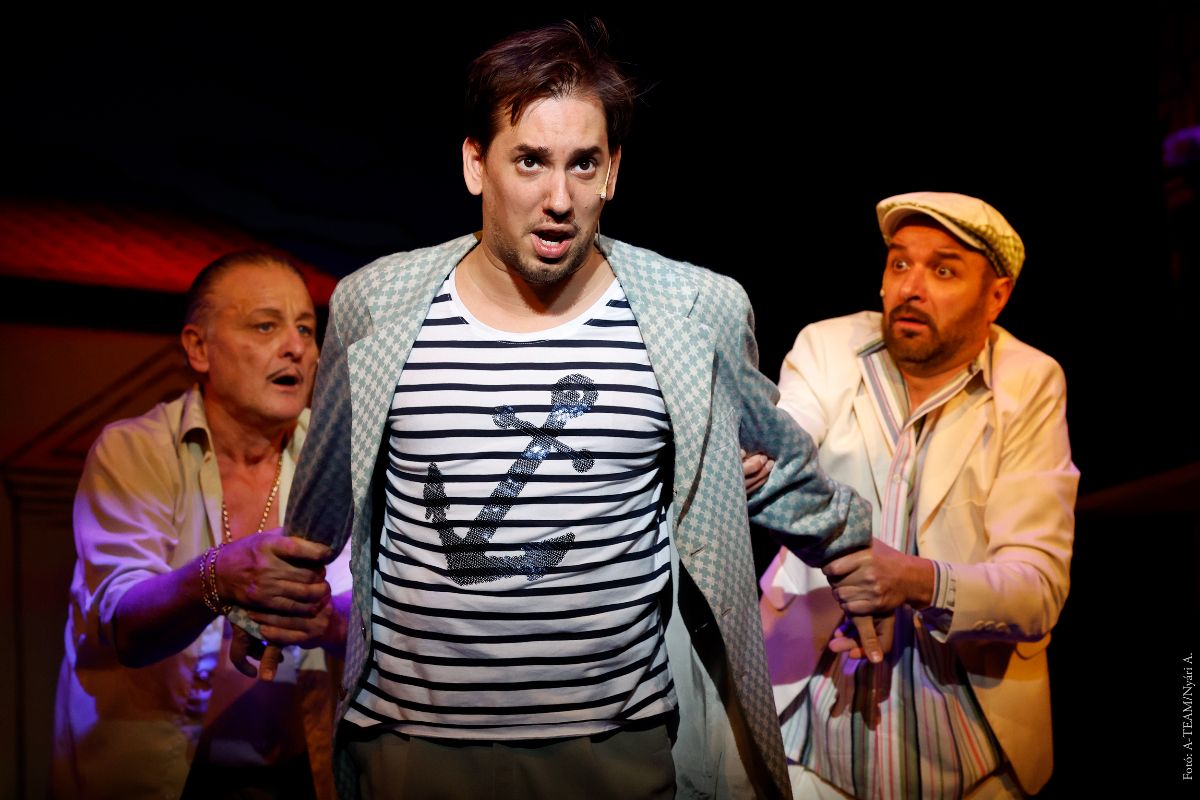 A színművészek az Anconai szerelmesek című előadásban jelenet közben a színpadon, miközben két férfi lefogja a harmadik szereplő karját.