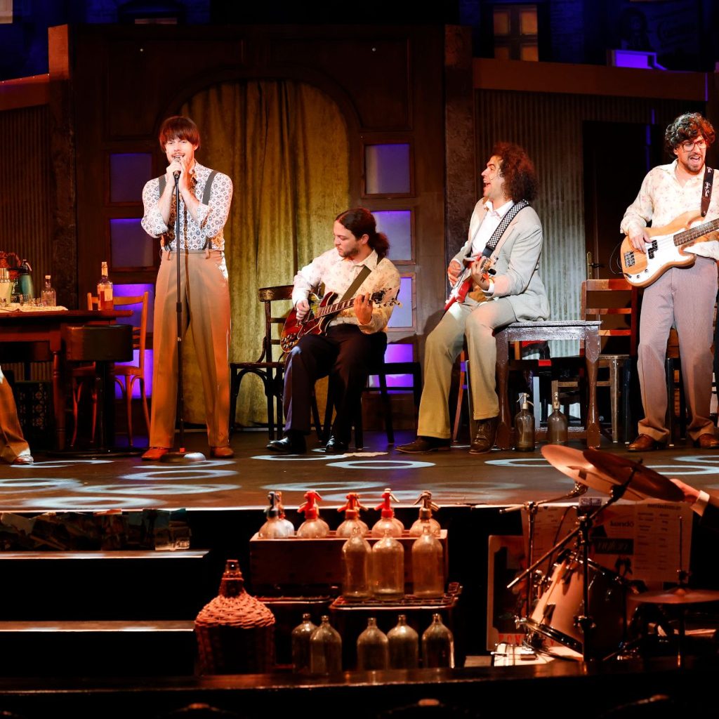 Azok a boldog szép napok című színházi előadás zenés jelenete, ahol a szereplők hangszereken játszanak és énekelnek a színpadon.