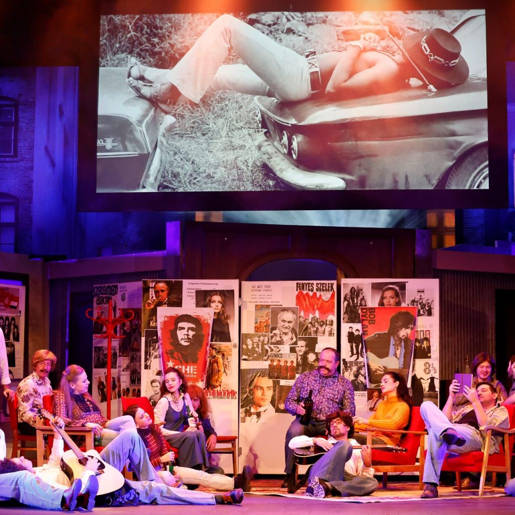 Azok a boldog szép napok című színházi előadás egyik jelenete, ahol a szereplők zenélnek és beszélgetnek a színpadon, a háttérben vetített fekete-fehér képpel.