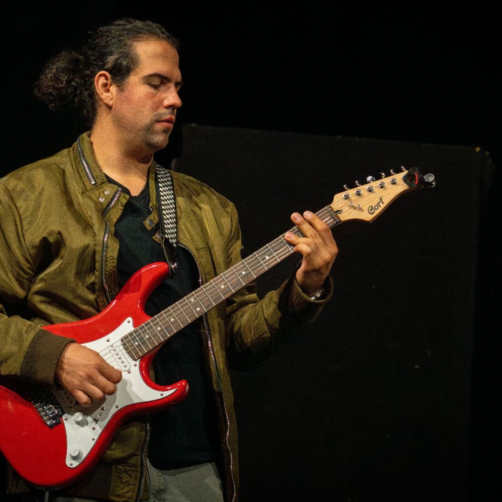 Egy hosszú, hátrakötött hajú férfi játszik egy piros elektromos gitáron a színpadon, barna kabátot visel, és sötét háttér előtt áll.