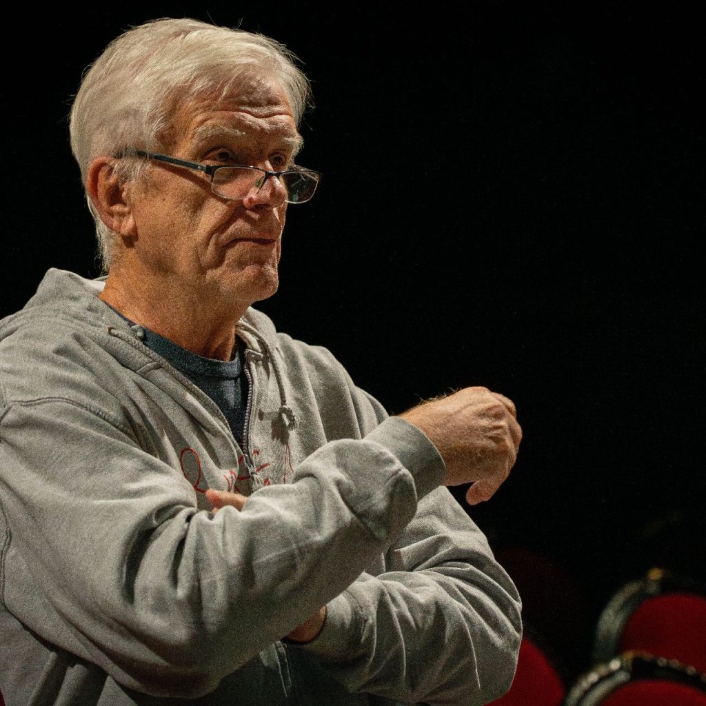 Szemüveges, szürke kapucnis pulóvert viselő férfi áll keresztbe tett karral, és úgy tűnik, hogy beszél vagy gesztikulál. A sötét háttérben piros székek láthatók.