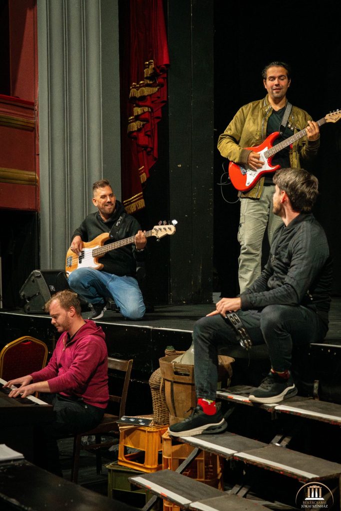 Négy férfi próbál a színpadon: egyikük elektromos gitáron, másikuk billentyűs hangszereken, harmadikuk basszusgitáron, harmadikuk pedig tamburát tart. Színházi kellékekkel és díszletekkel vannak körülvéve, mosolyognak és interakcióba lépnek egymással.