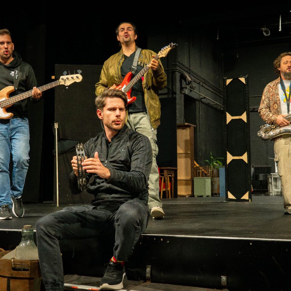 Négy férfi lép fel egy színpadon: egy basszusgitáron játszik, egy a peremén ülve tamburával énekel, egy arany kabátban áll, egy pedig szaxofonozik. A háttér egy fekete, minimalista színházi díszlet.