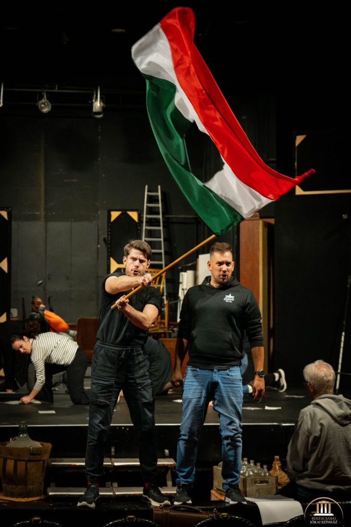 Két férfi áll a színpadon; az egyik egy a magyar zászlót lenget. A háttérben más emberek és színpadi kellékek láthatók.