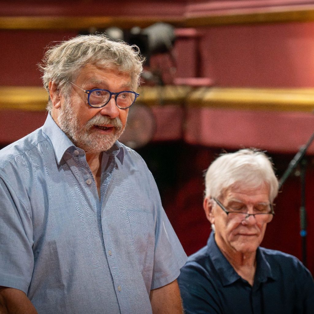 Két ősz hajú, szemüveges, idősebb férfi van bent. Az egyik áll és beszél, a másik ül és lefelé néz. Egy színházteremben vannak, amelynek falai vörösek és aranyszínűek.