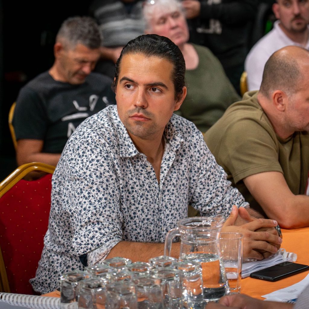 Egy férfi mintás ingben ül egy asztalnál, üvegkancsókkal és papírokkal, és úgy tűnik, mintha egy megbeszélés vagy megbeszélés közben figyelné.