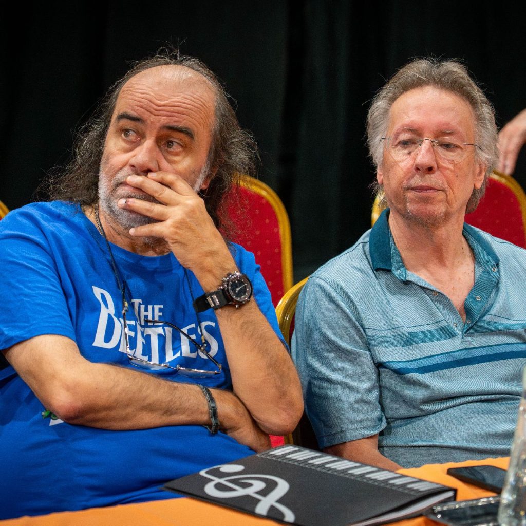 Két férfi ül egymás mellett egy piros székekből álló asztalnál. Az egyik férfi kék Beatles-inget visel, és kezét az arcán pihenteti; a másik csíkos inget visel. Az asztalon egy mappa, egy vizes kancsó és poharak állnak.