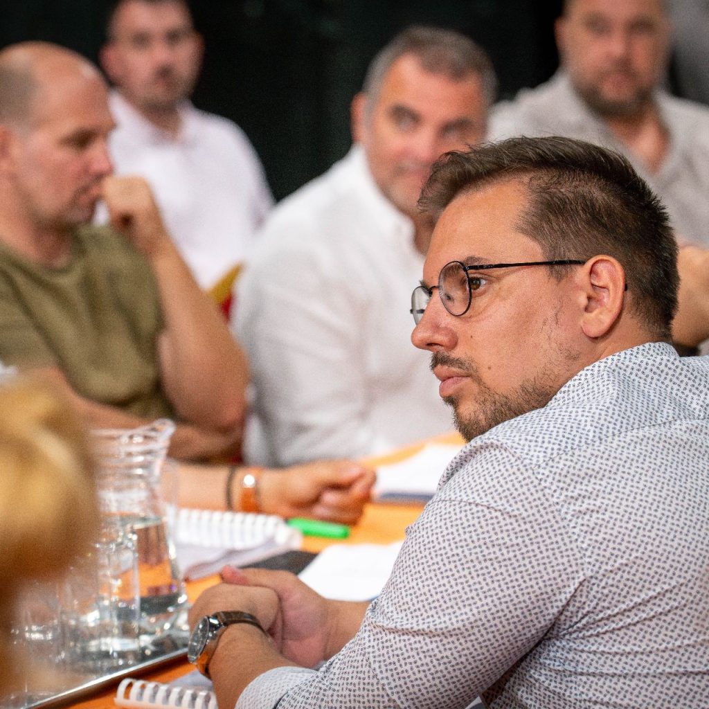 Emberek egy csoportja ül egy asztal körül, és beszélgetésbe elegyedik. Egy szemüveges, fehér inges férfi figyelmesen hallgatja. Az asztalon jegyzetfüzetek, papírok és vizes kancsók állnak. Úgy tűnik, hogy a helyszín egy megbeszélés vagy konferencia.