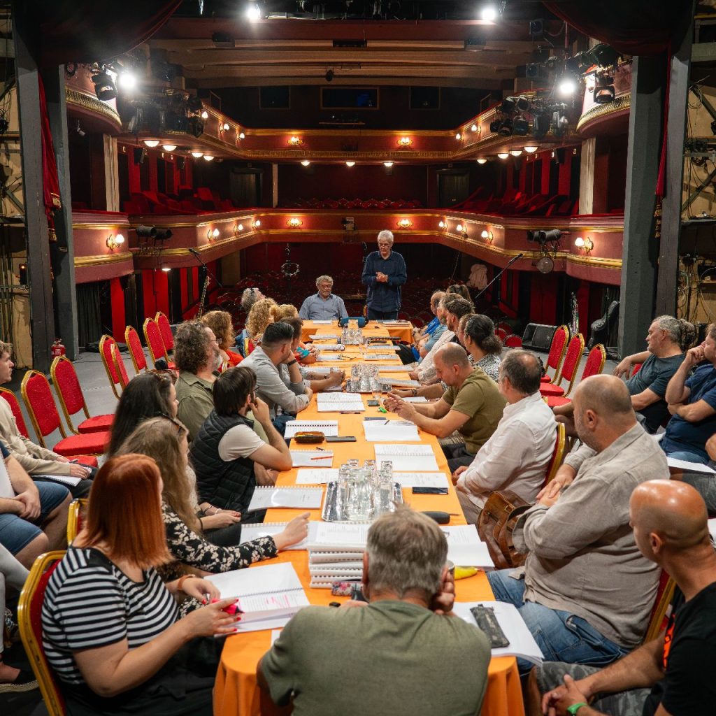 Egy színházi színpadon egy nagy téglalap alakú asztal körül ül egy csoport ember, akik egy megbeszélésen vagy próbán vesznek részt, előttük papírok és forgatókönyvek. A háttérben a színház piros székei és erkélye látható.
