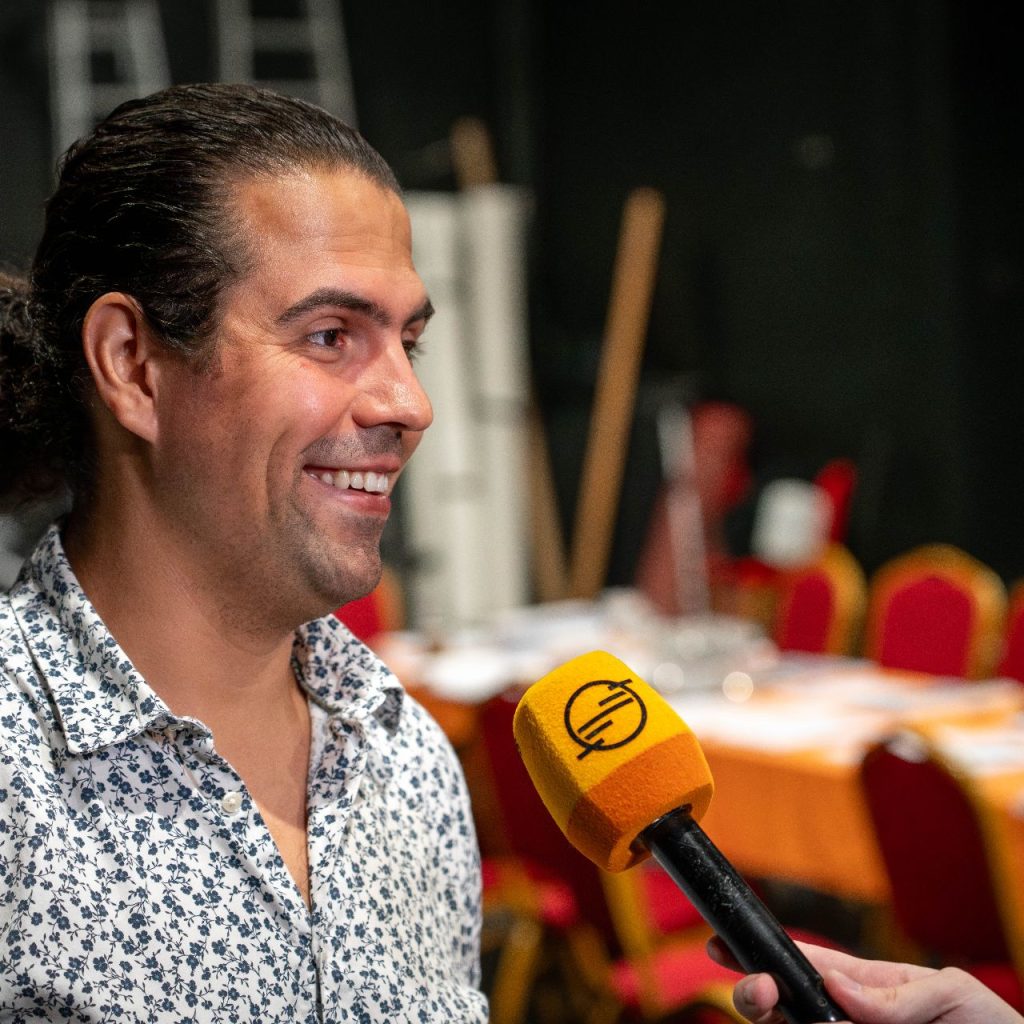 Egy hosszú, sötét hajú, mintás inges, mosolygós férfit kérdeznek sárga mikrofonnal. Mögötte piros székek, egy narancssárga terítővel letakart asztal és elmosódott backstage tárgyak.