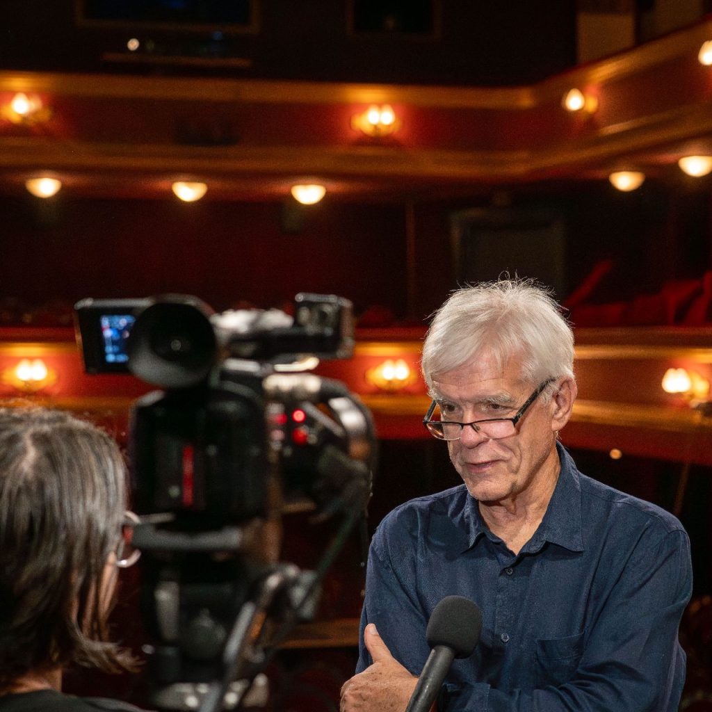 Egy idősebb, fehér hajú férfi áll egy színházban, és egy interjú során mikrofonba beszél, miközben az előtérben kamera és interjúkészítő látható.