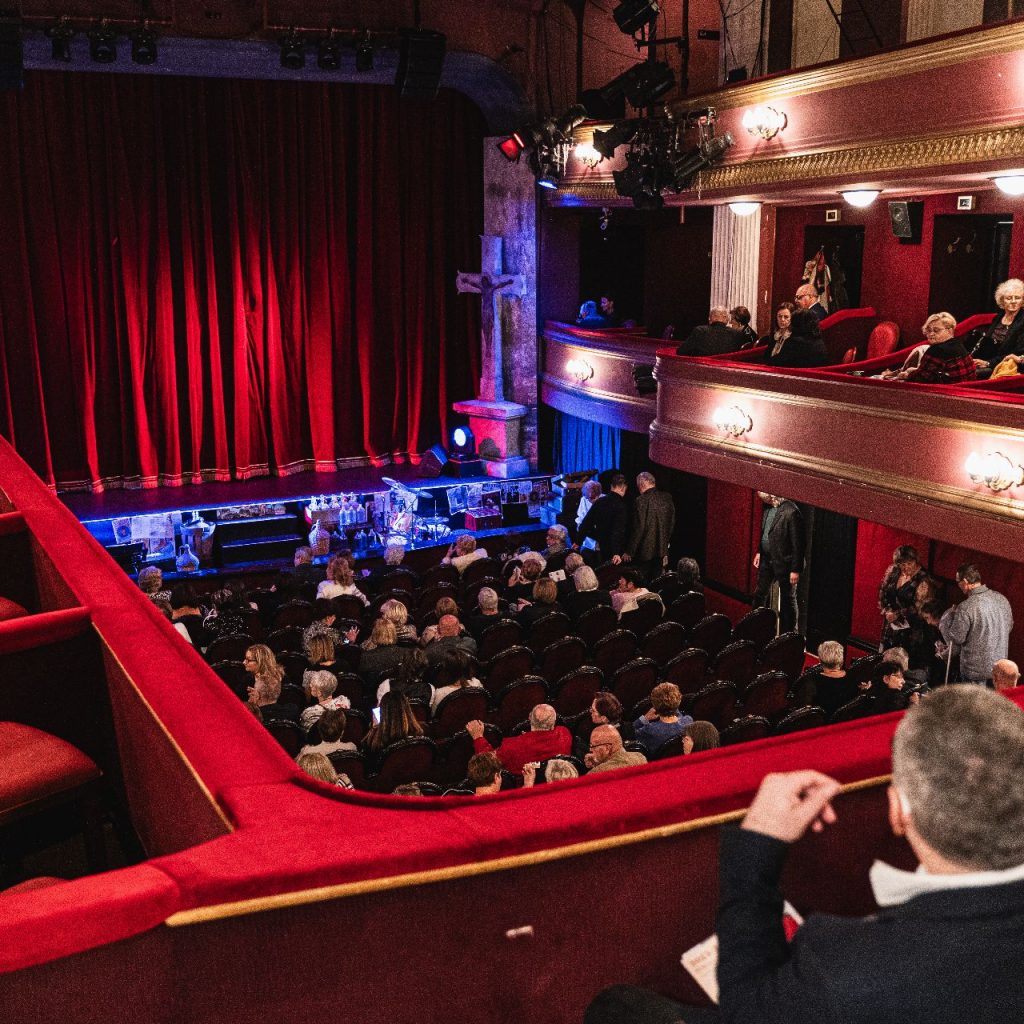 A nézők egy piros ülésekkel és aranyszínű díszítéssel díszített színházban ülnek és beszélgetnek, és várják az előadást. A színpadon zárt vörös függöny és halvány világítás van. Néhányan a színpad felé néznek.