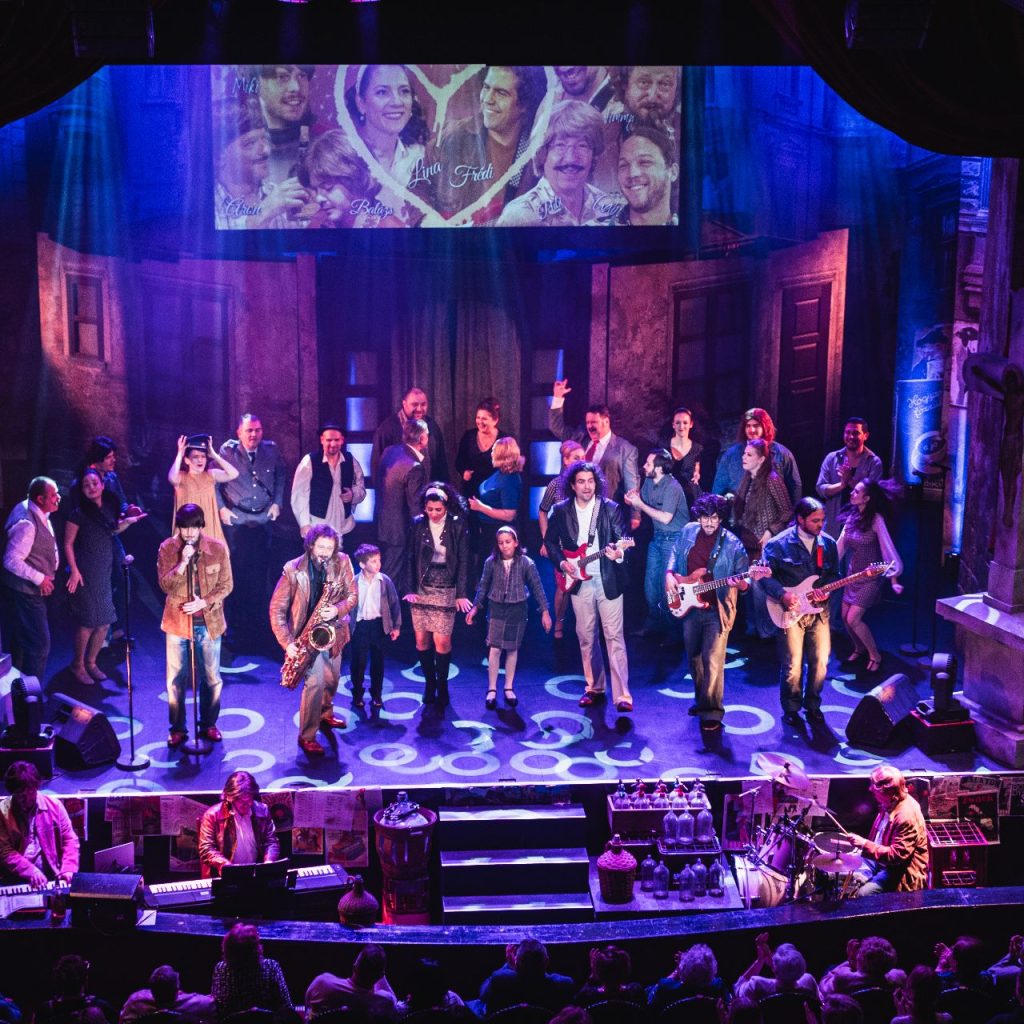 Egy nagyszámú szereplő énekel és játszik hangszereken egy vibráló színházi színpadon, színes világítással, hangszerekkel és a föléjük vetített különböző emberek képével. A közönség alulról nézi a produkciót.