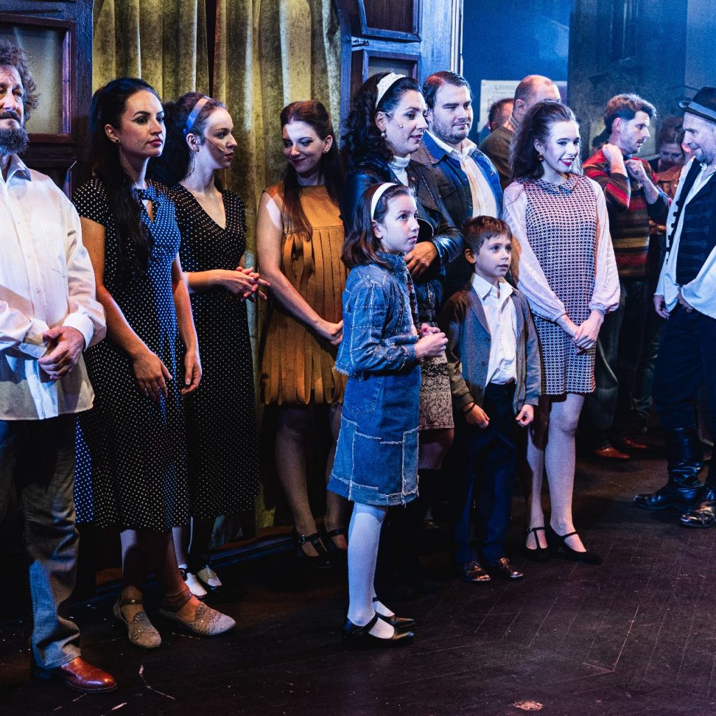 A színészek egy csoportja, köztük felnőttek és gyerekek, együtt állnak a színfalak mögött, jelmezben. Néhányan előre néznek, míg mások beszélgetnek. A színpadi fények megvilágítják őket, a háttérben pedig színpadi kellékek és függönyök láthatók.