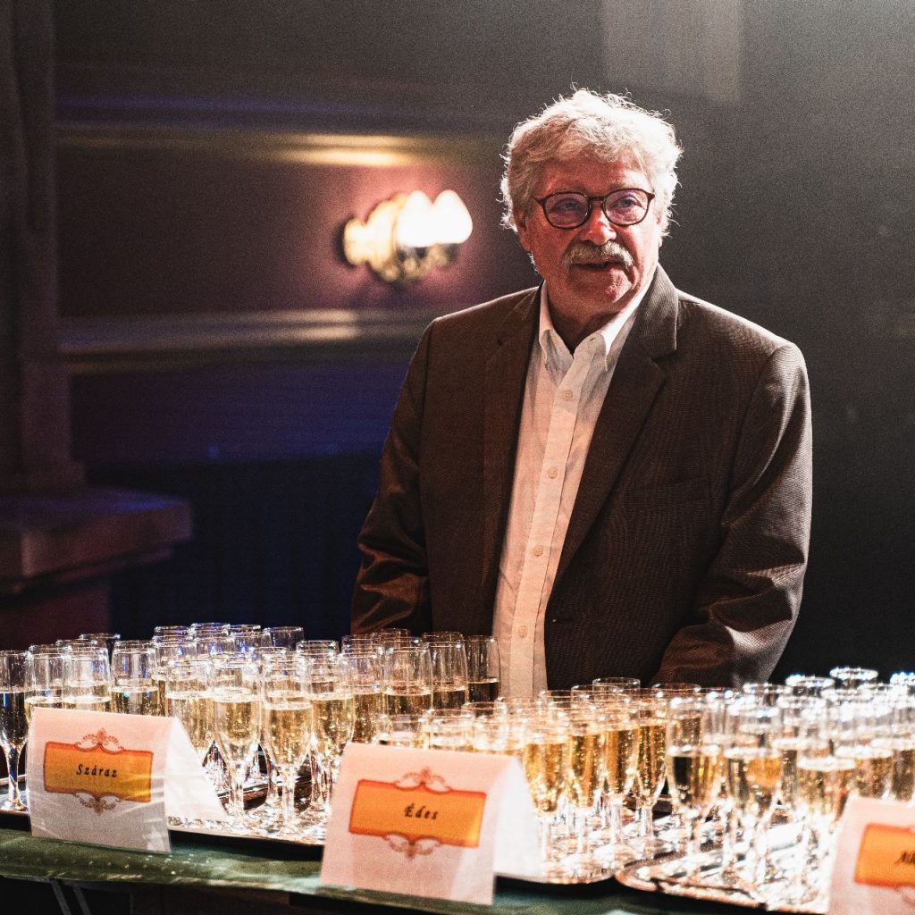 Egy öltönyös, ősz hajú, szemüveges, idősebb férfi áll egy pezsgőspoharakkal teli asztal mögött egy beltéri rendezvényen. A meleg világítás ünnepi hangulatot teremt.