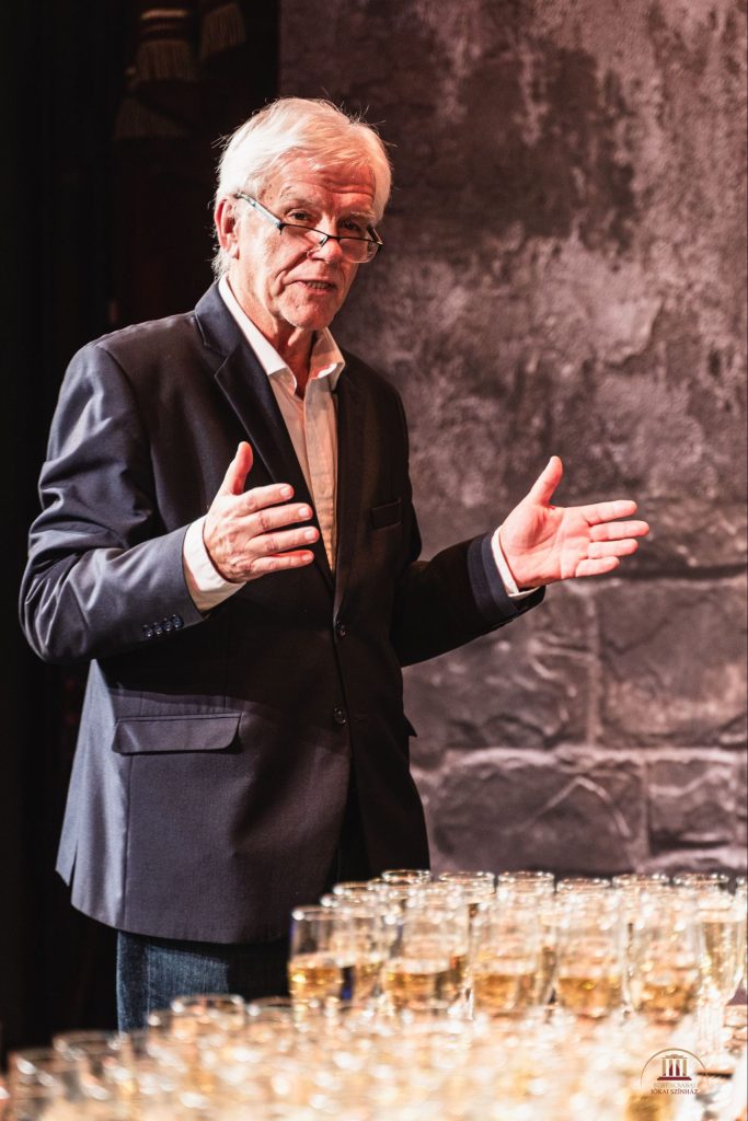 Egy idősebb, ősz hajú, szemüveges, sötét öltönyt és fehér inget viselő férfi gesztikulál a kezével, miközben egy asztal mellett áll, amely tele van sok pohár itallal. A háttérben egy texturált kőfal.