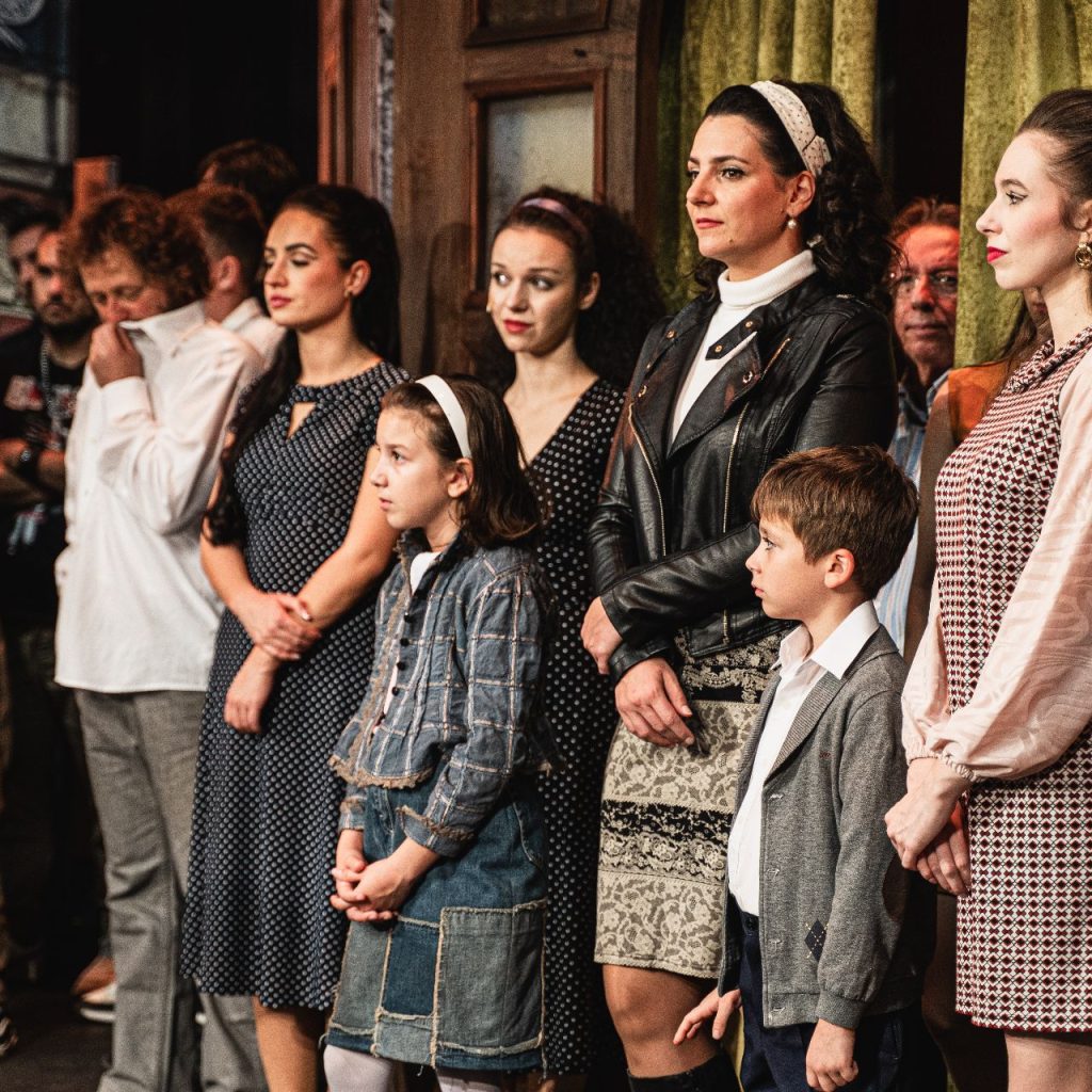 Emberek egy csoportja, köztük felnőttek és gyerekek, fél- és alkalmi öltözékben, előrefelé fordulva, figyelmes arckifejezéssel, mintha egy előadást vagy eseményt néznének.