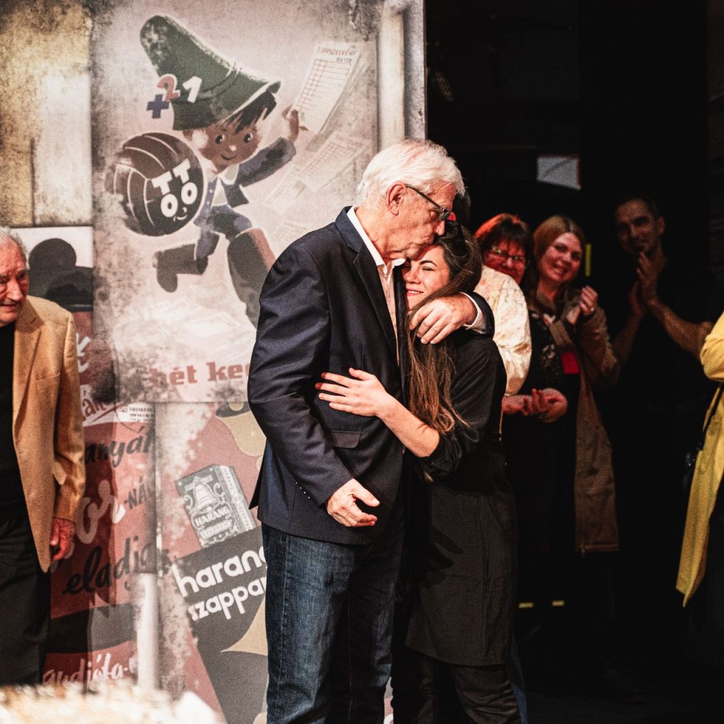 Egy blézeres férfi megölel egy nőt a színpadon, miközben az emberek körülöttük állnak, tapsolnak és mosolyognak. Mögöttük egy színes plakát látható egy rajzfilmfigurával. A jelenet melegnek és ünnepélyesnek tűnik.