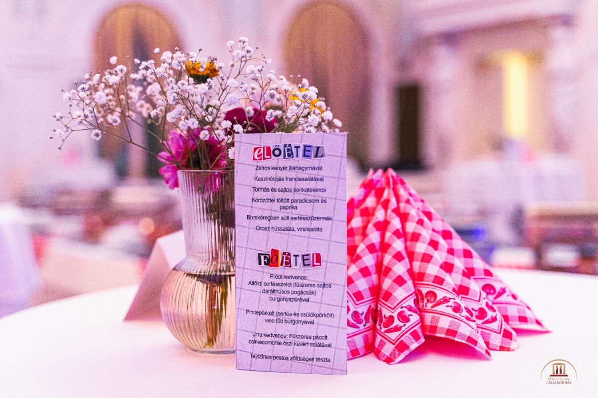 Virágos asztaldísz és piros kockás szalvéta egy fehér terítőn, mellettük menükártya ’Előétel’ és ’Főétel’ felirattal, elegáns rendezvény helyszínén.