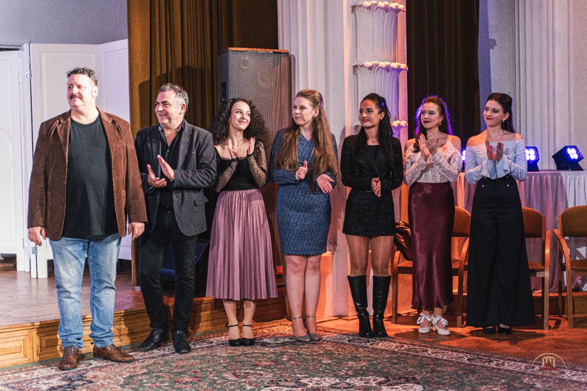 Egy elegáns teremben nyolc ember áll egymás mellett, mosolyogva és tapsolva. A nők ünnepi ruhát viselnek, a férfiak zakóban állnak a színpad peremén, mögöttük oszlopok és függönyök láthatók.