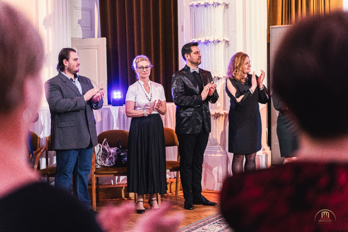 Felnőttek sora áll és tapsol egy elegáns teremben. A háttérben oszlopok, barna függönyök és székek láthatók, az előtérben nézők alakjai homályosan kirajzolódnak.