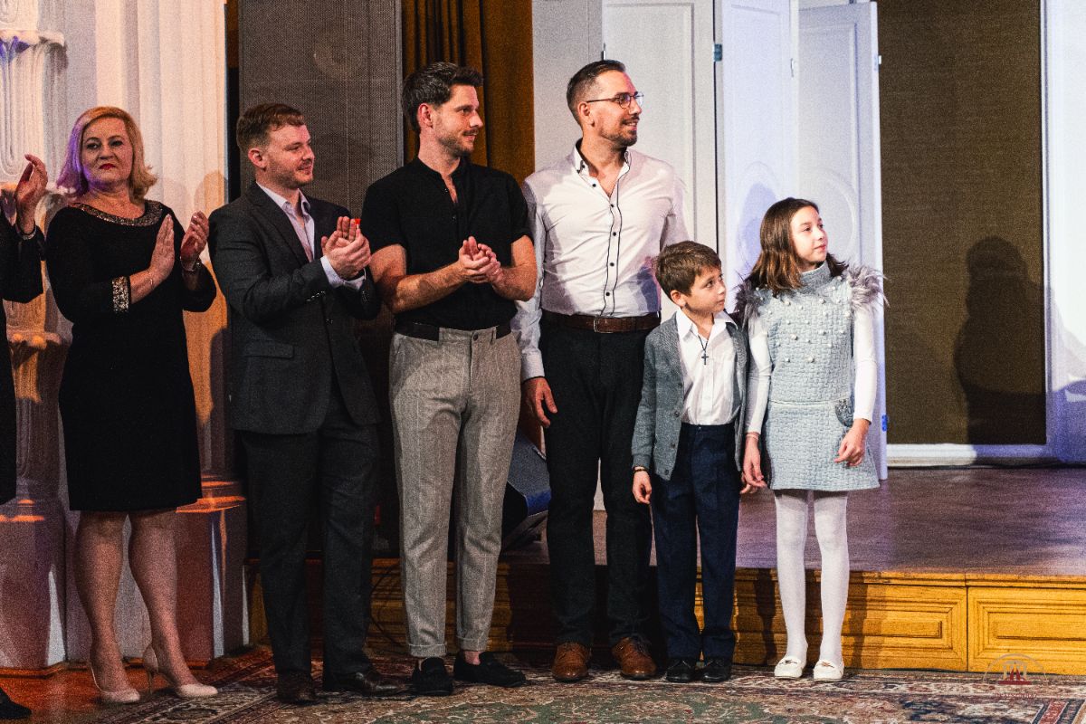 Színpadon álló felnőttek és két gyermek elegáns ruhában, egymás mellett tapsolnak és mosolyognak. A háttérben világos ajtók és barna függöny látható, a hangulat ünnepélyes.