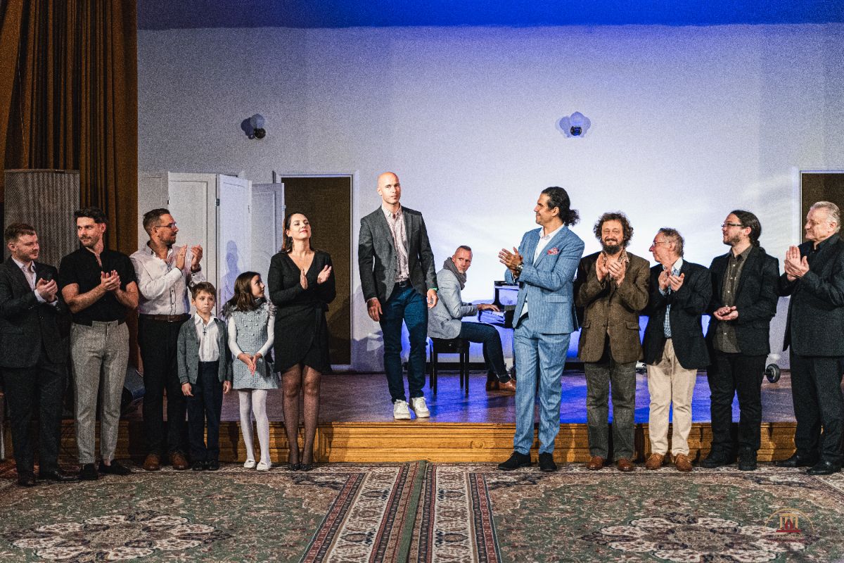 Színpadon álló emberek tapsolnak, miközben középen egy férfi épp előlép. Mellette elegánsan öltözött nők, férfiak és gyerekek állnak, a háttérben egy zongorista játszik.