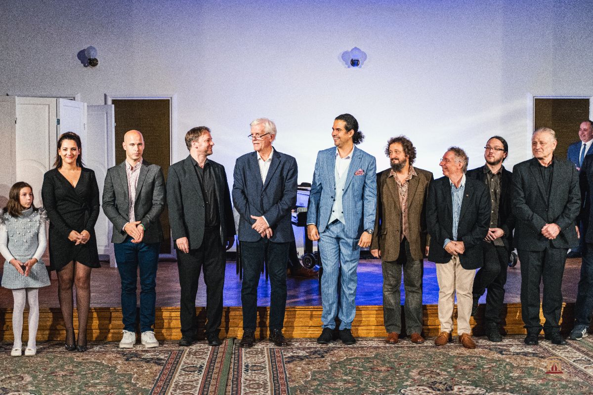 Tizenegy ember áll egymás mellett egy rendezvényen, mosolyogva néznek egymásra. Elegánsan öltözött férfiak és nők, köztük egy kislány is, szőnyeggel borított emelvényen, kék háttérfény előtt.