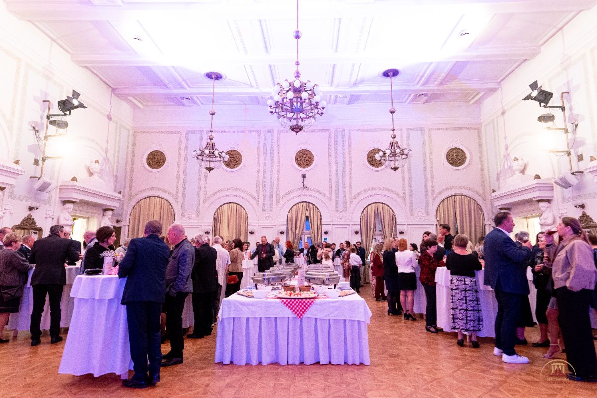 Elegáns bálterem, ahol vendégek állnak és beszélgetnek a svédasztalos vacsora körül. A díszes mennyezetről csillárok lógnak, a háttérben arany függönyök és klasszikus faldíszek láthatók.