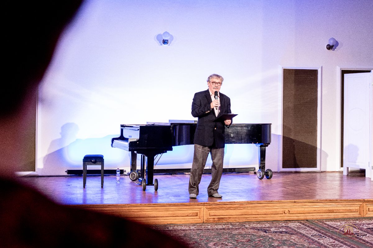 Ősz hajú férfi zakóban mikrofont tart és beszédet mond a színpadon, mögötte egy fekete zongora látható. A háttér világos, meleg fényekkel megvilágított, a jelenet bensőséges hangulatot áraszt.