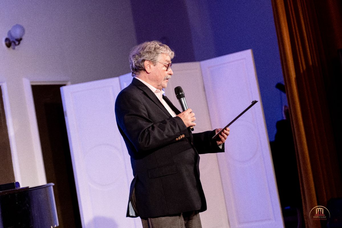 Ősz hajú férfi zakóban mikrofont tart és papírról olvas a színpadon. A háttérben világos paravánok és függönyök láthatók, a megvilágítás meleg, bensőséges hangulatot teremt.