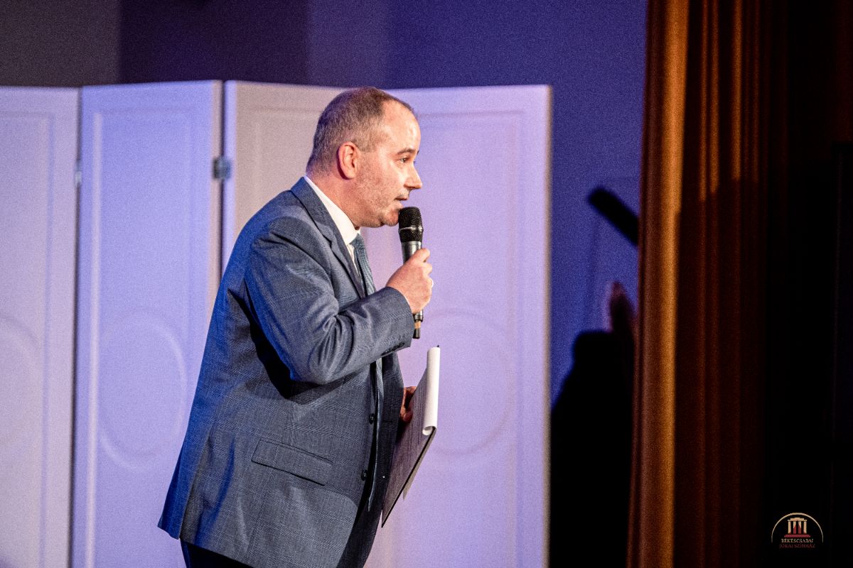 Férfi elegáns öltönyben mikrofont tart a kezében és beszédet mond a színpadon. A háttérben fehér paravánok és barna függöny láthatók, a fények meleg hangulatot teremtenek.