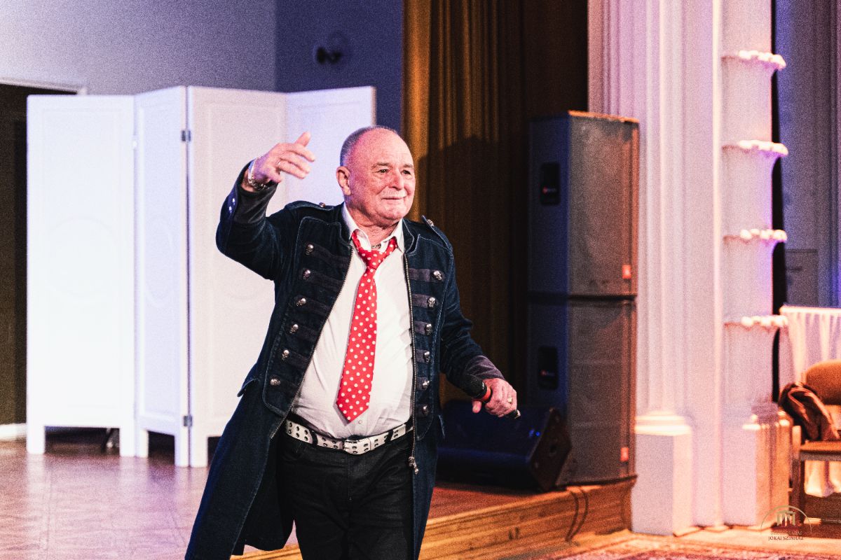 Idősebb férfi mosolyogva áll a színpadon, elegáns díszruhát és piros pöttyös nyakkendőt visel. A háttérben oszlopok, hangfalak és barna függönyök láthatók, a hangulat ünnepi.