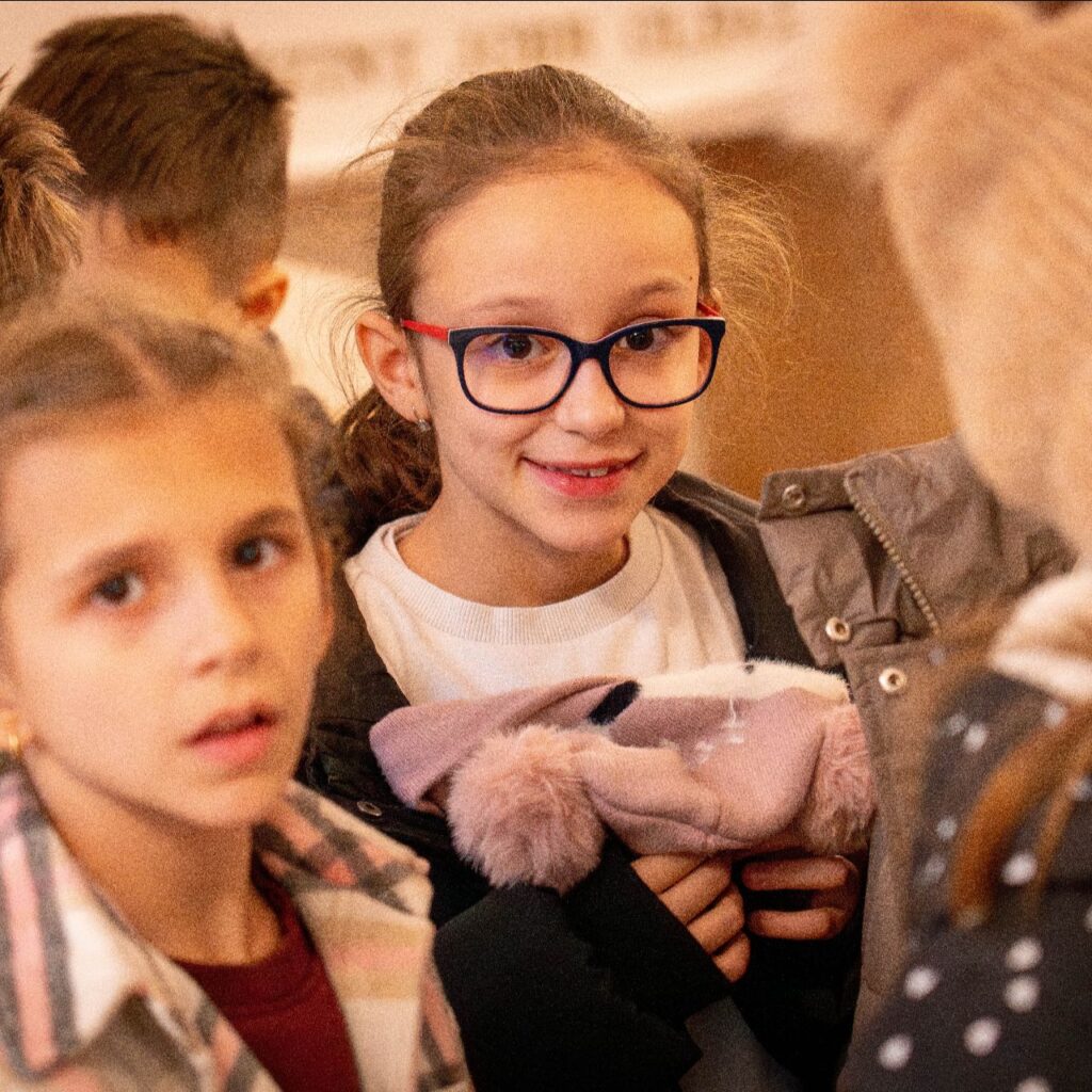 Gyerekek vannak a képen, középen a legélesebb részen egy szemüveges kislány mosolyog a kamerába