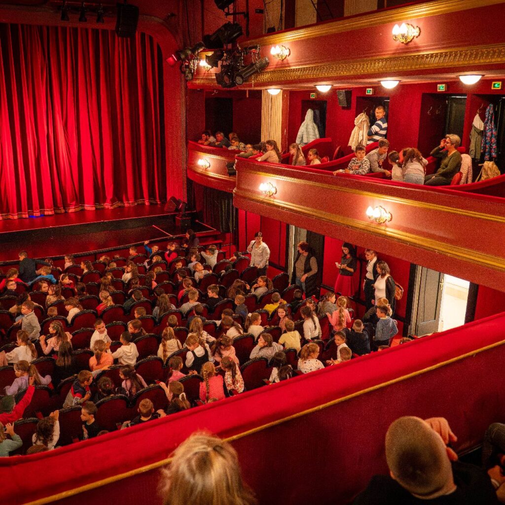 Bal oldalt látható a színpad, leengedett vörös függönyökkel, a képen lent pedig a nézőközönség van, akik mind gyerekek. Páholyból lett fotózva