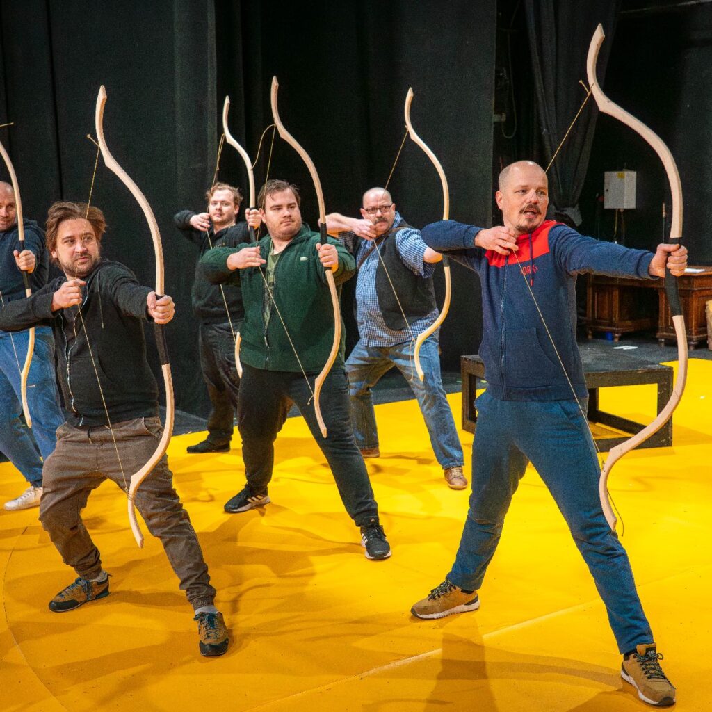 Öt férfi a színpadon áll, íjat fognak a kezükben, jobbra fordulnak és éppen kihúzzák az íjat.