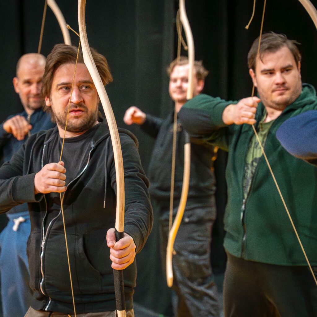 Öt férfi a színpadon áll, íjat fognak a kezükben, jobbra fordulnak és éppen kihúzzák az íjat.