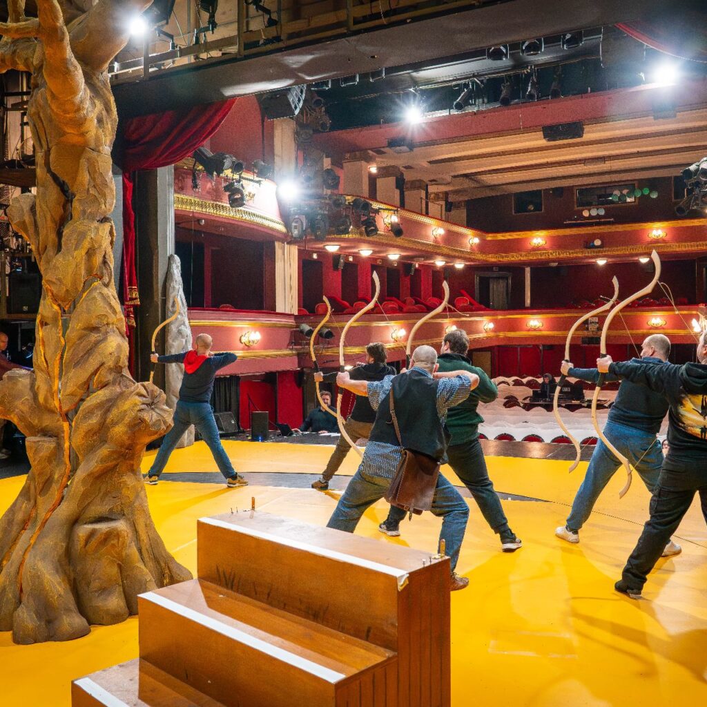 Öt férfi a színpadon áll háttal, íjat fognak a kezükben, balra fordulnak és éppen kihúzzák az íj húrját, mintha lőnének. Az előtérben egy nagy díszlet fa van, a háttérben pedig a páholyok.