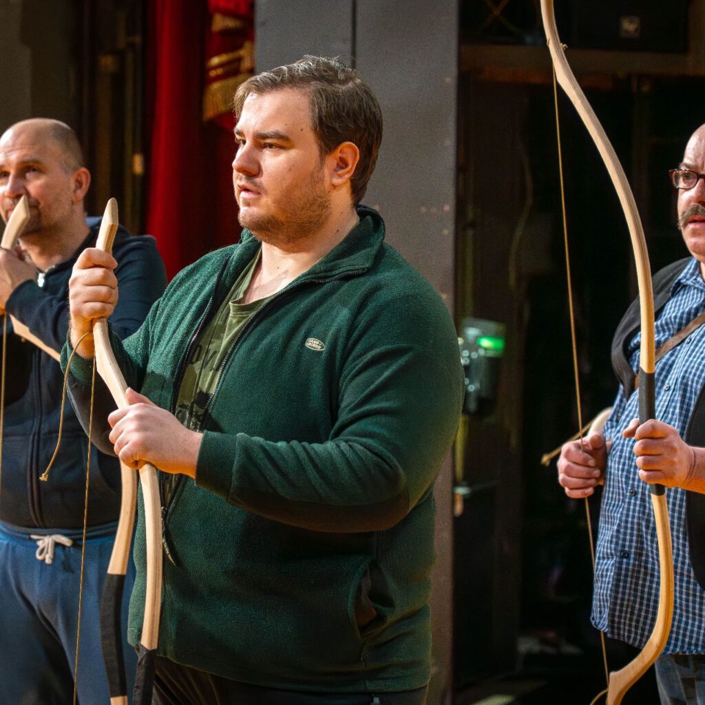 Hátorm férfi áll, a kezükben tartják az íjat, azon támaszkodnak és figyelik ahogyan a rendező beszél.