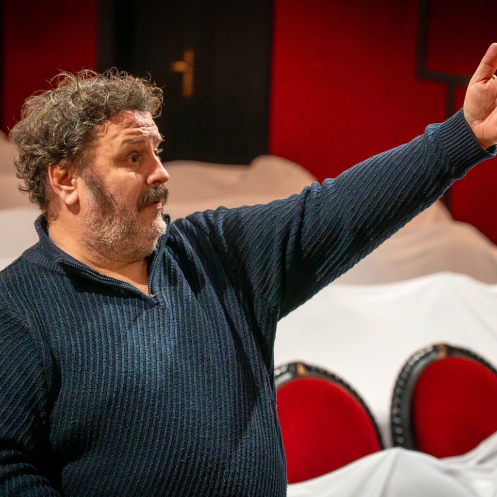 A Rege a csodaszarvasról rendezője Csurulya Csongor áll a színpad előtt, és a színpad irányába mutatva beszél.