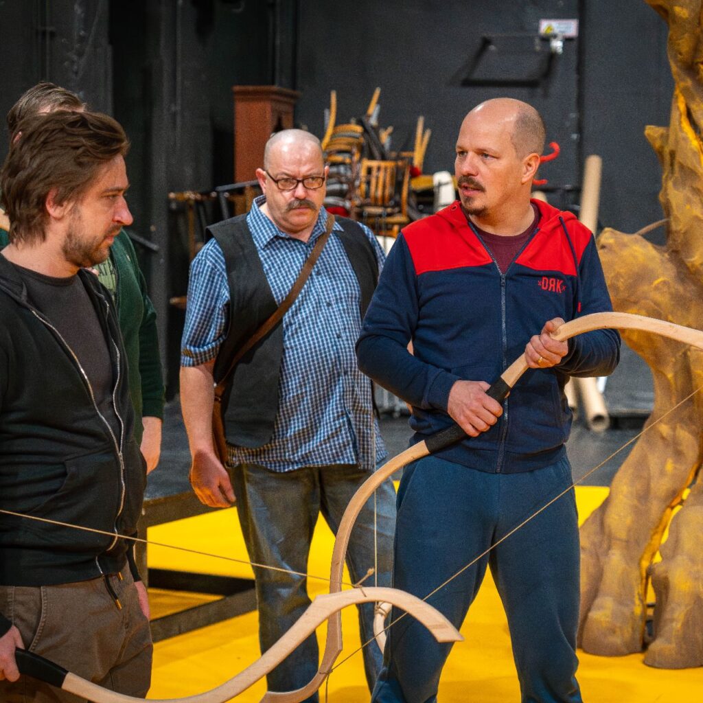 Négy férfi áll a színpadon és beszélgetnek. A háttérben jobb oldalt egy díszlet elem van, ami fatörzset ábrázol.