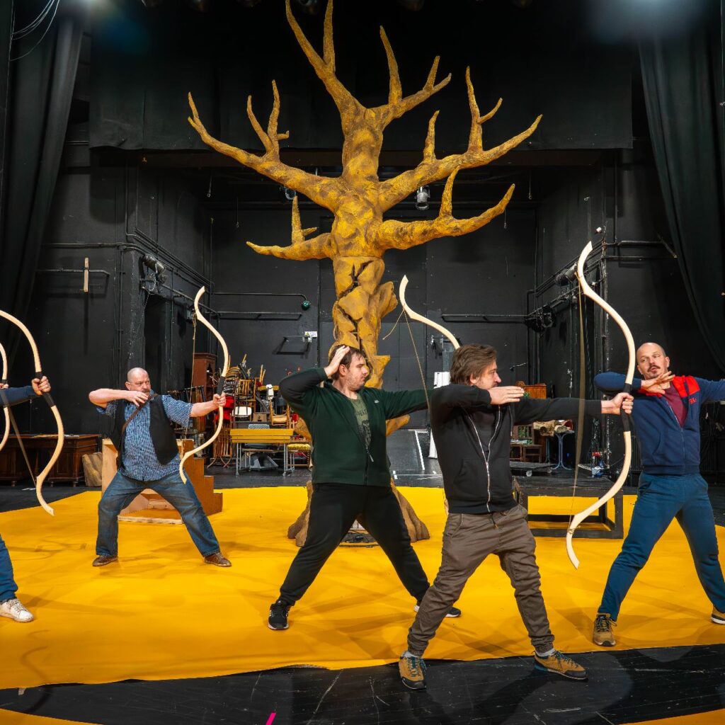 Öt férfi a színpadon áll, íjat fognak a kezükben, jobbra fordulnak és éppen elengedték az íj húrját, mintha lőnének. A háttérben egy nagy díszlet fa van.