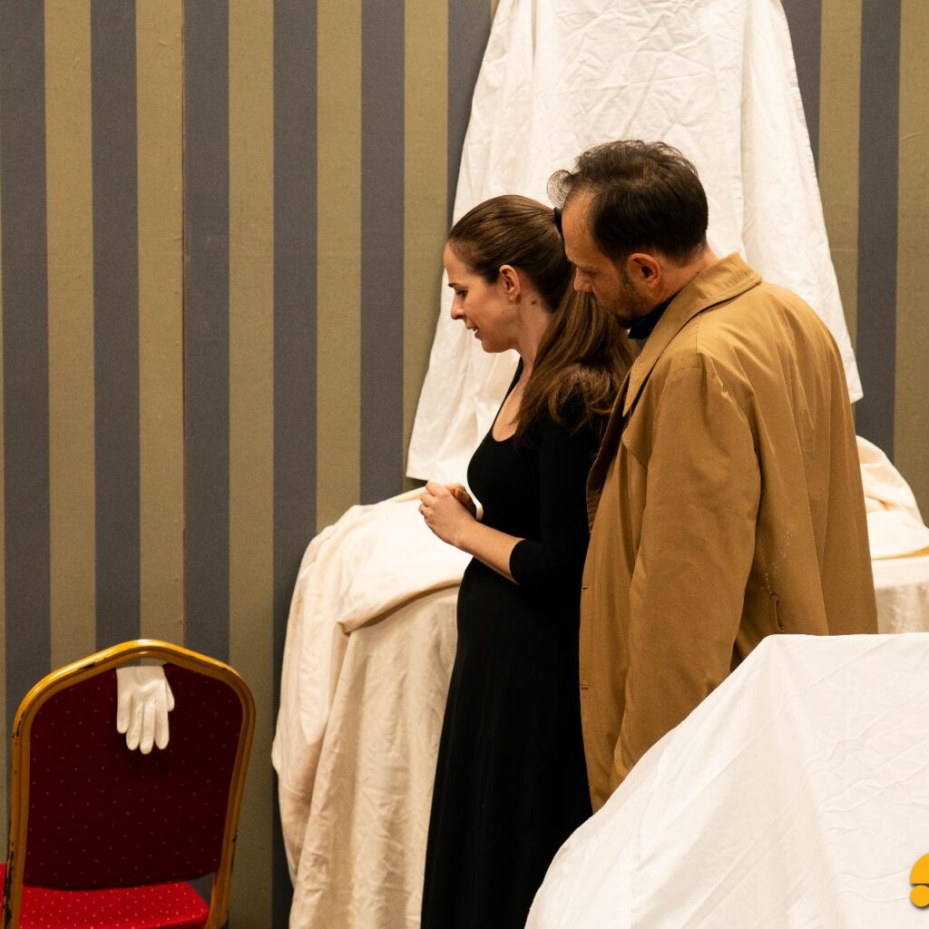 Egy nő fekete ruhában és egy férfi barna kabátban egymás mellett áll, bal oldalra néznek egy széken lévő kesztyűre