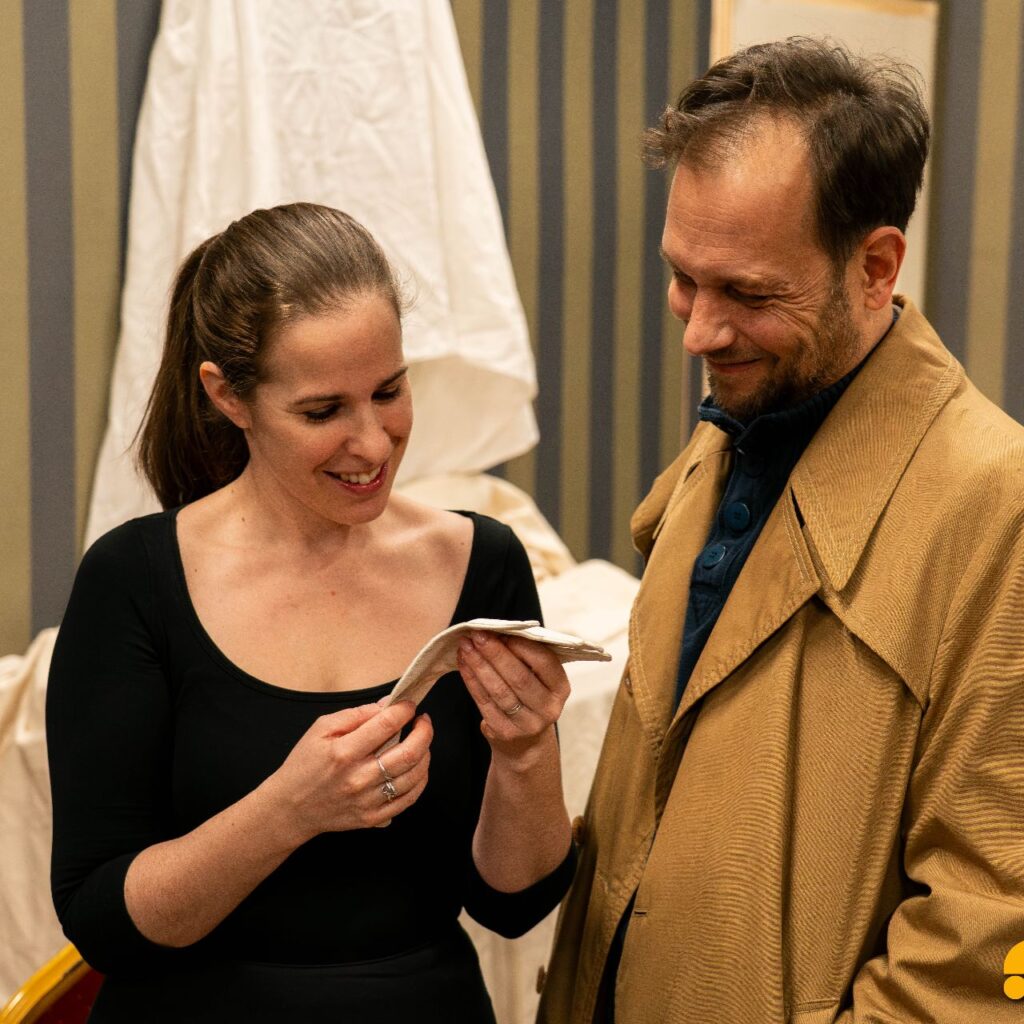 Egy nő fekete ruhában és egy férfi barna kabátban egymás mellett áll, mosolyogva néznek a nő kezében tartott fehér kesztyűre