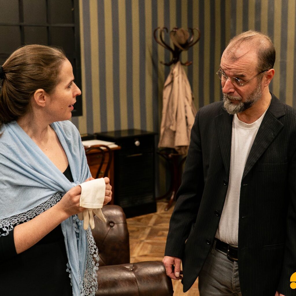 Bal oldalt egy nő áll összefogott hajjal, a hátán kék kendő van, a kezében egy fehér kesztyűt tart, és jobbra néz, ahol egy férfi áll fekete zakóban, kör szemüvege van, és éppen a nőnek mond valamit