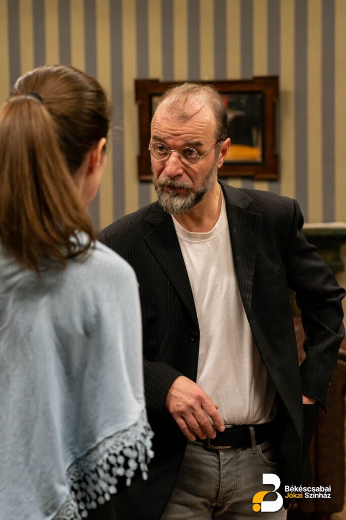 Az előtérben egy nő látható háttal, össze van fogva a haja és kék kendő van a hátán. A fókuszban egy férfi van fekete zakóban és kerek szemüveggel. A férfi éppen a nőre néz és beszél