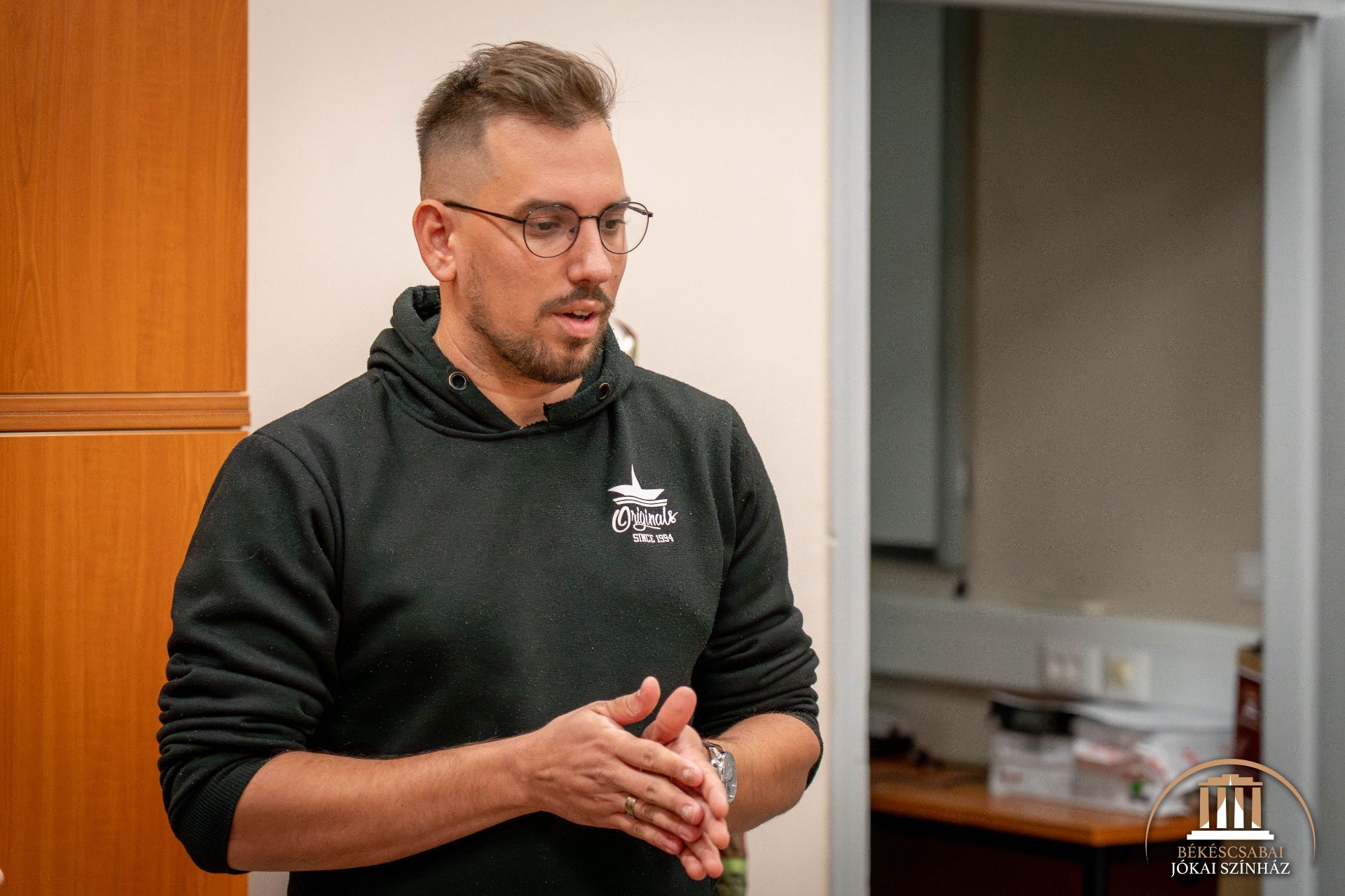 Egy rövid hajú, szemüveges, rövidre nyírt szakállú, fekete kapucnis pulóvert viselő férfi áll bent, és a kezével gesztikulál. Balra tőle egy szekrény, a háttérben pedig egy íróasztal irodai tárgyakkal.