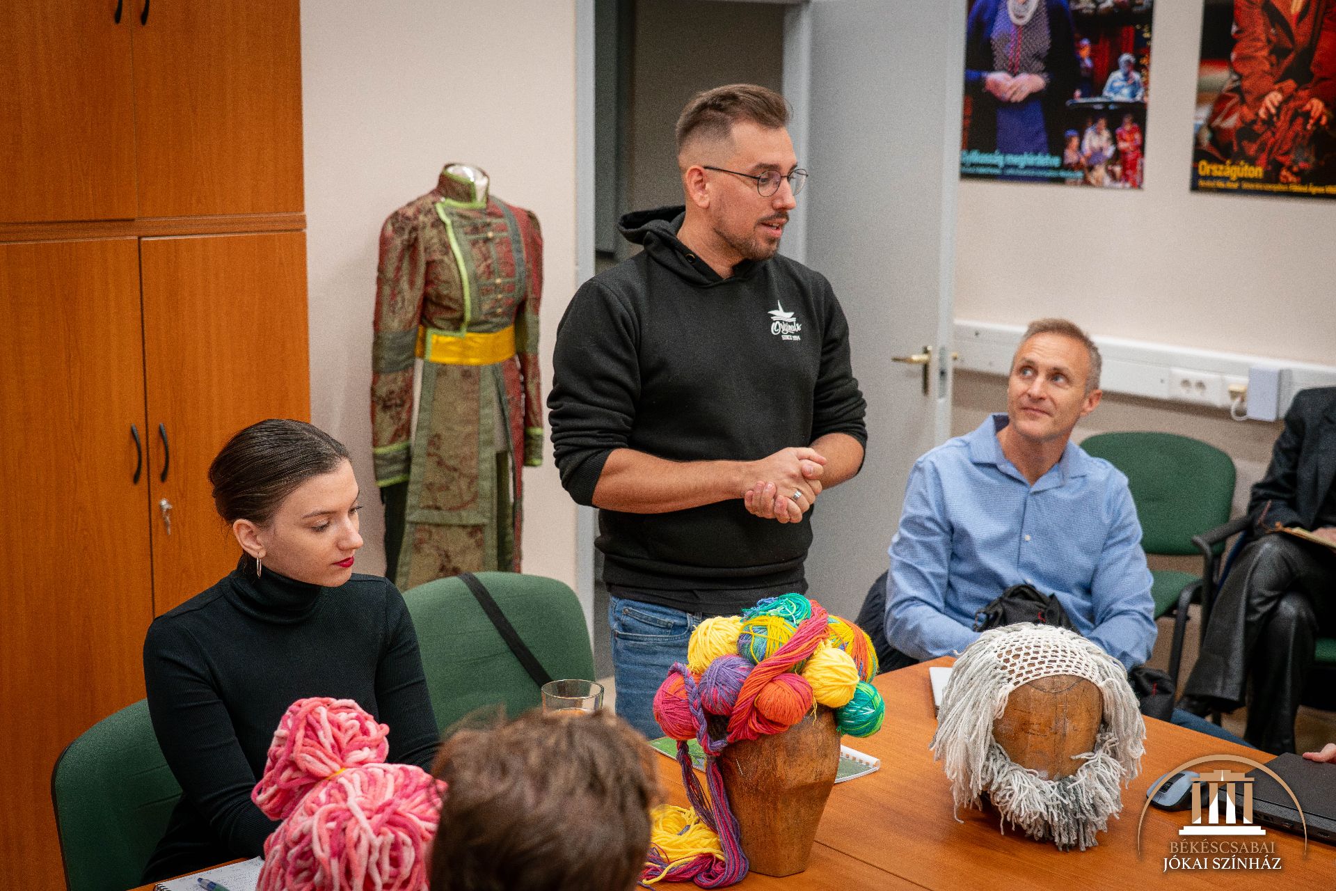 Egy férfi áll és beszél ülő kollégáihoz egy színházi jelmezekkel és plakátokkal díszített teremben. Az előttük lévő asztalon színes fonalak és parókák láthatók.