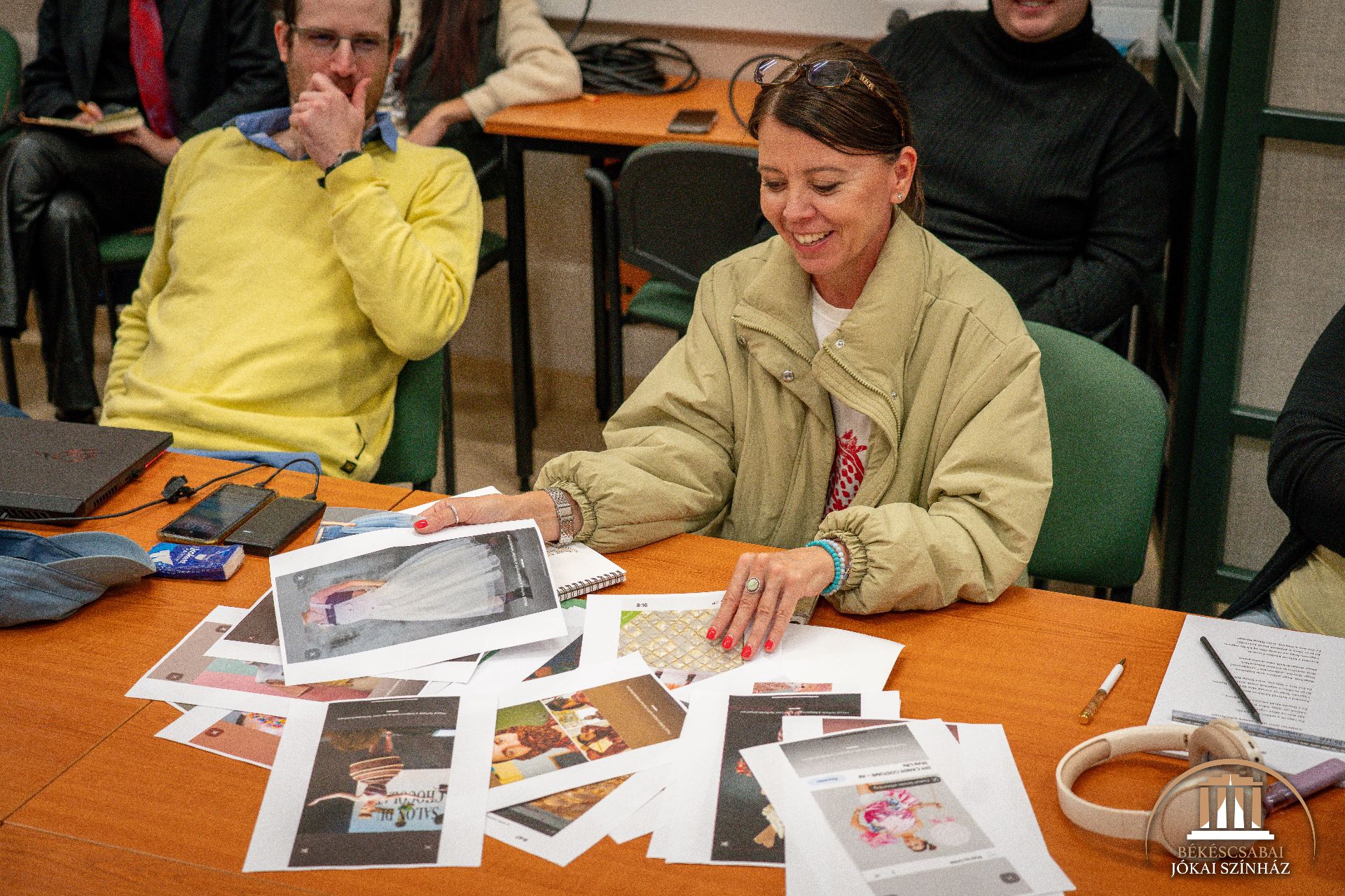 Egy bézs kabátos nő mosolyogva nézi végig az asztalon kiterített, kinyomtatott fényképeket. A nő körül mások is ülnek, és a légkör laza és együttműködőnek tűnik.