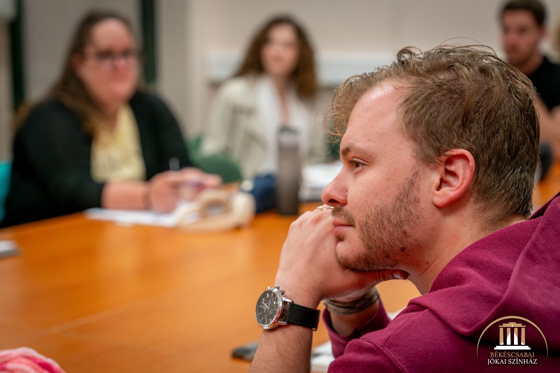 Egy piros kapucnis férfi ül egy asztalnál, állát a kezére támasztva, elgondolkodó tekintettel. A háttérben elmosódva más emberek láthatók, akik látszólag megbeszélésen vagy megbeszélésen vesznek részt. A jobb alsó sarokban egy logó látható.